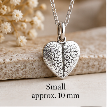 A silver heart-shaped pendant necklace with a textured, crackled leaf-like pattern, hanging on a delicate silver chain. The pendant is displayed on a stone slab next to dried white flowers, with text overlay at the bottom reading 'Small approx.