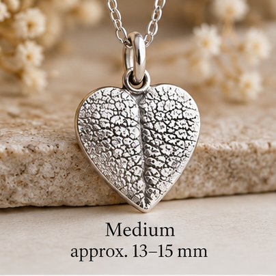 A silver heart-shaped pendant necklace with a textured leaf-like pattern, hanging from a delicate silver chain. The pendant is displayed against a soft beige background with dried flowers and a stone slab, with text overlay reading 'Medium approx.