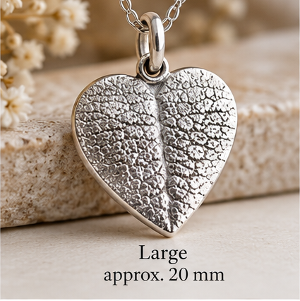 A silver heart-shaped pendant with a textured leaf-like pattern on a matching chain, displayed on a stone surface with dried flowers in the background. Text at the bottom reads 'Large approx.