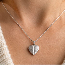 A close-up of a person wearing a delicate silver chain necklace with a small textured heart pendant, paired with a cream-colored knit top.