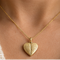 A close-up of a person wearing a delicate gold chain necklace with a textured, heart-shaped pendant, styled with a cream-colored knit top.