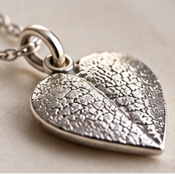 A silver heart-shaped pendant necklace with a textured, leaf-like surface detail, hanging from a silver chain on a neutral beige background.