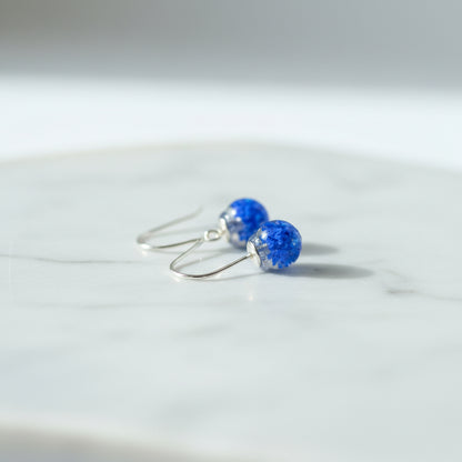 Blue earrings with a floral design held in a hand against a neutral background