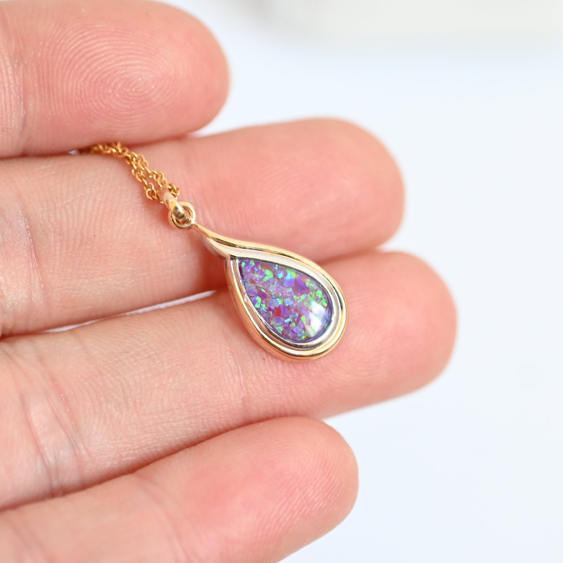 Gold pendant with a colorful stone held between fingers on a white background