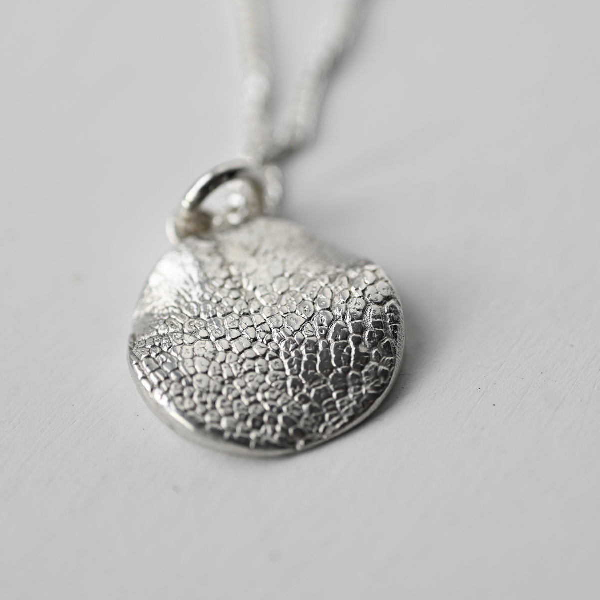 A textured silver pendant necklace with a hammered, organic surface pattern on a matching silver chain, photographed on a neutral light gray background.