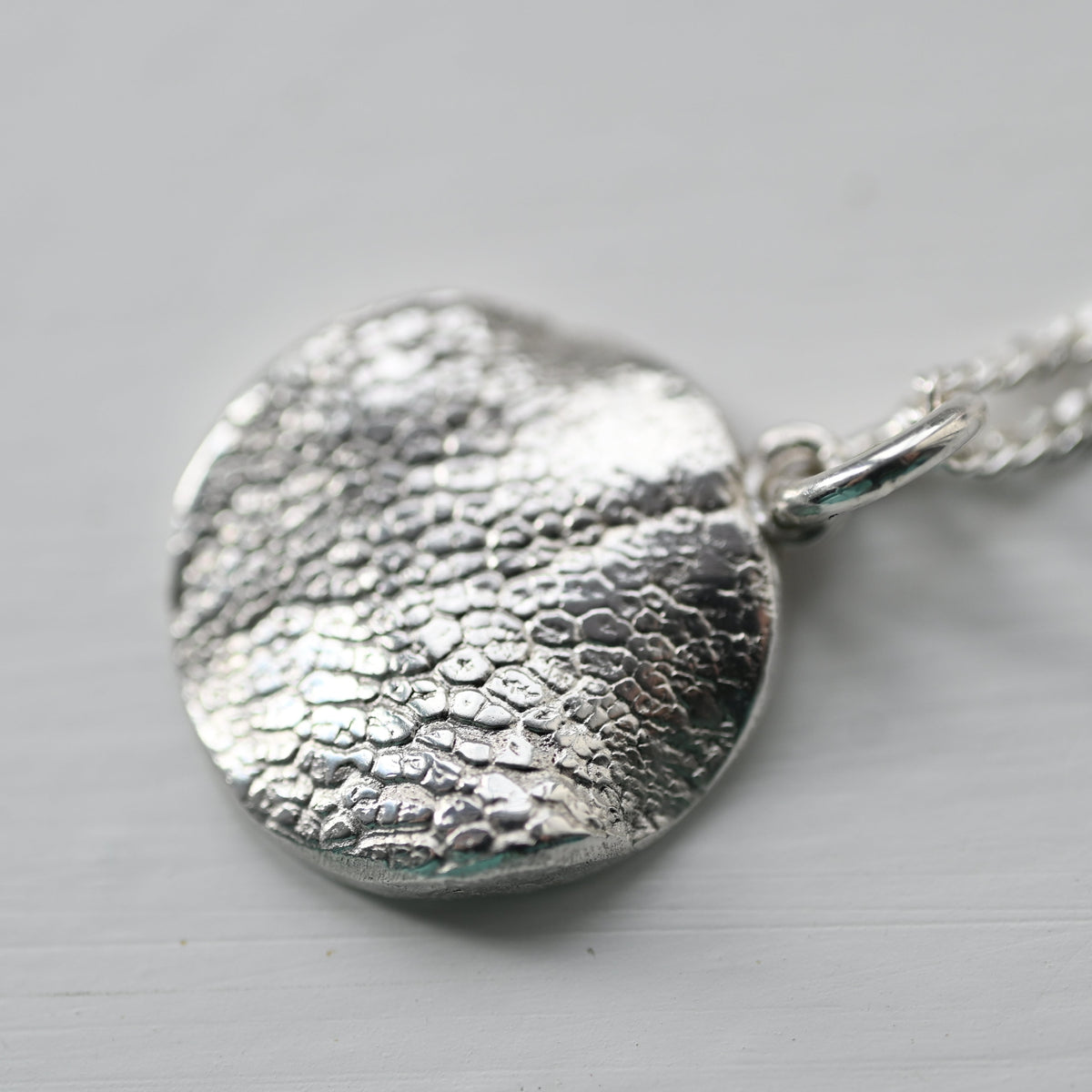A textured sterling silver pendant necklace with a hammered, organic surface pattern on a matching silver chain, shown on a light gray background.
