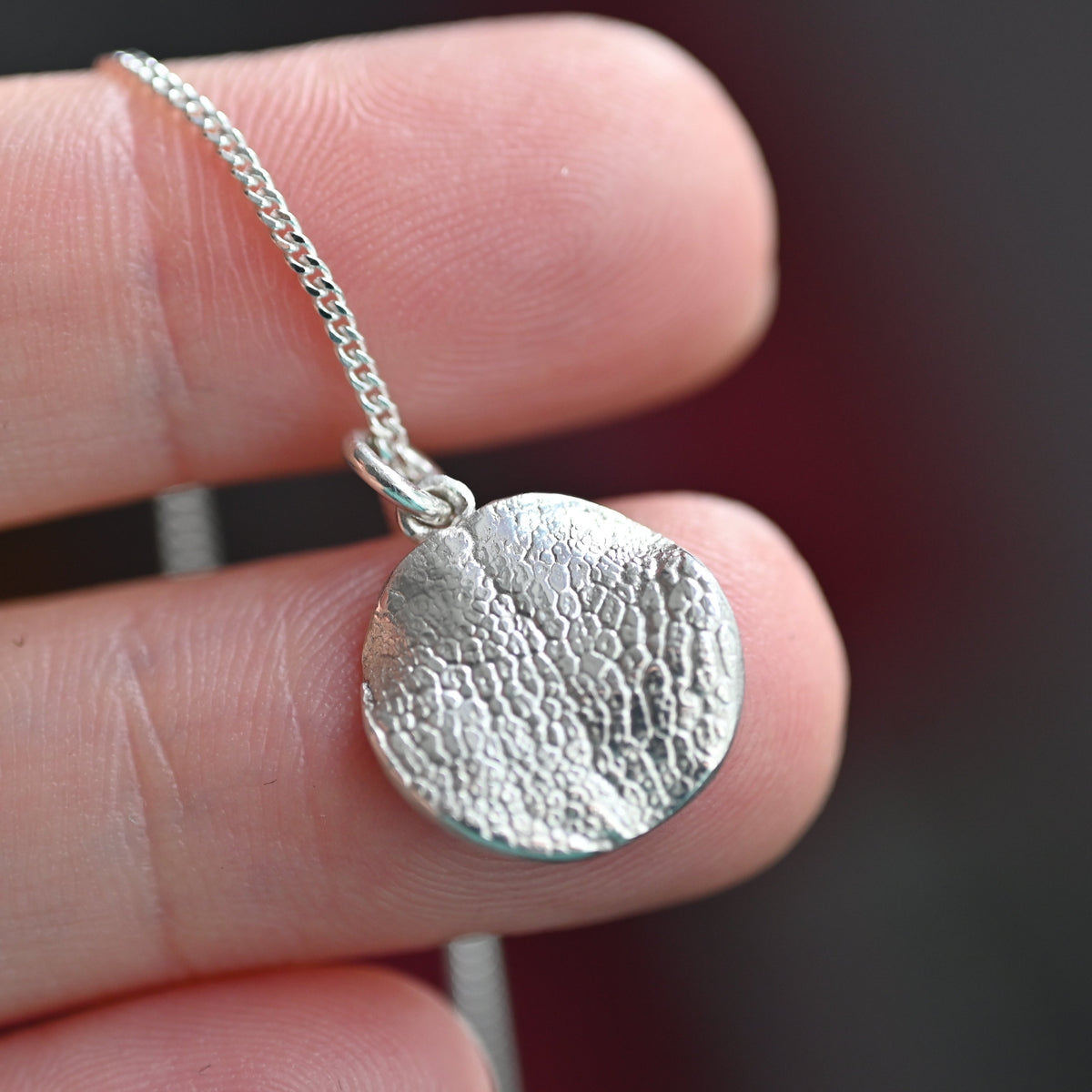 A close-up of a hand holding a small textured silver pendant necklace on a delicate chain against a dark blurred background.