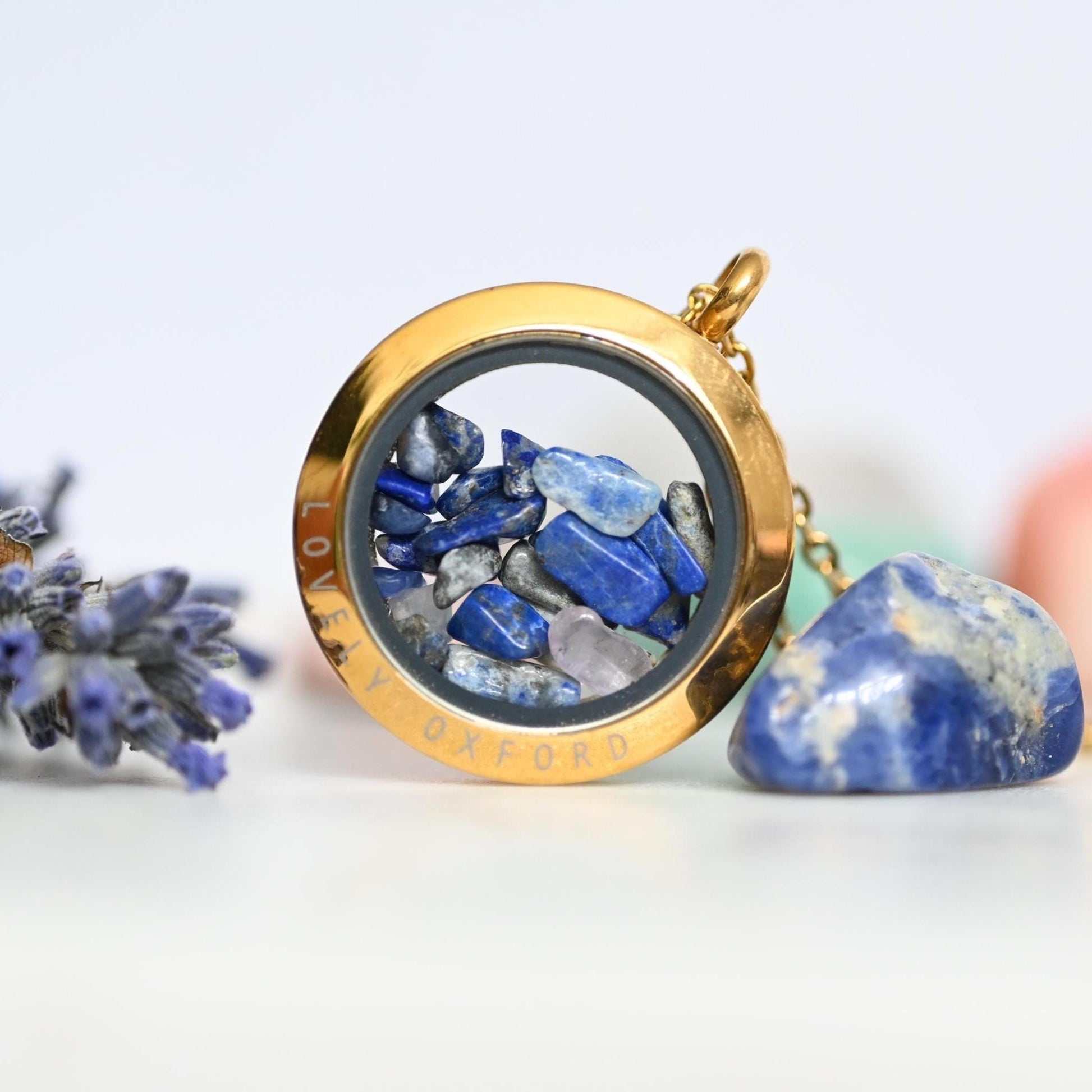 Gold locket with blue stones and 'Lovely Oxford' text, surrounded by lavender flowers on a light background