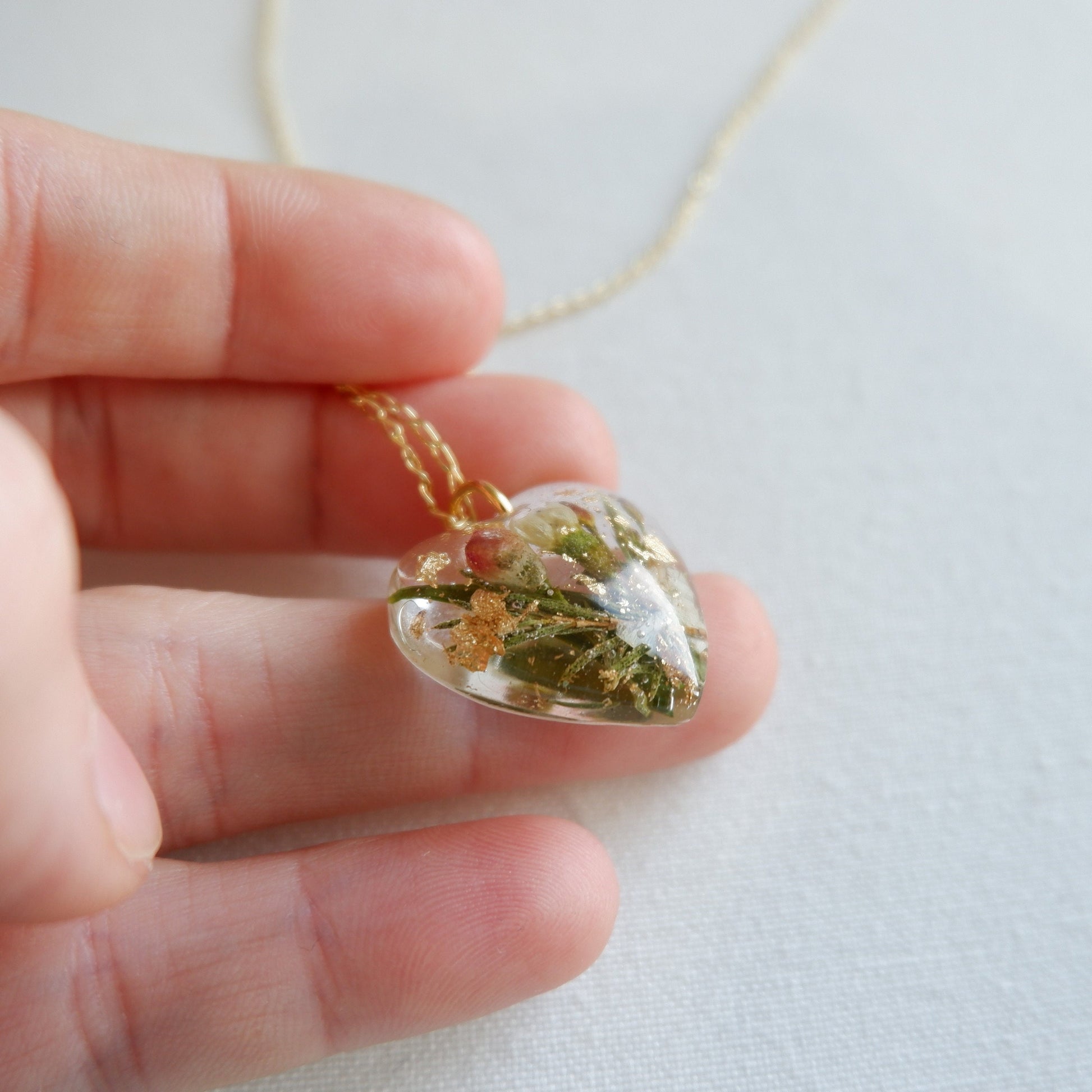 Heart-shaped pendant with greenery held in a hand against a light background