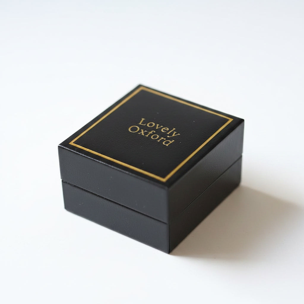 A black square jewelry box with gold foil borders and the brand name 'Lovely Oxford' printed in gold on the lid, photographed on a white surface.
