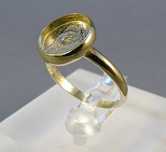 A gold ring with a textured, circular bezel setting, displayed on a clear plastic stand against a gray background.