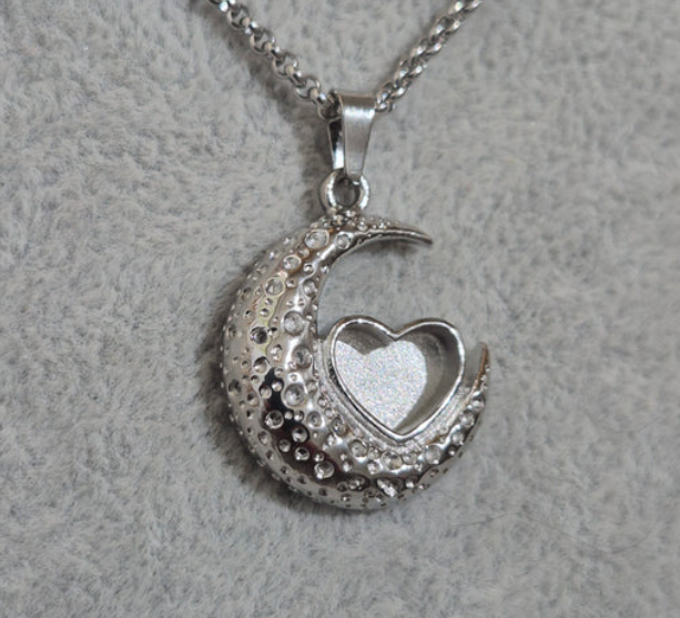 A silver crescent moon pendant necklace with a small open heart-shaped locket in the center, decorated with clear rhinestones, shown on a textured gray background.