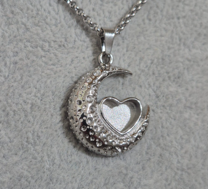 A silver crescent moon pendant necklace with a small open heart-shaped locket in the center, decorated with clear rhinestones, shown on a textured gray background.