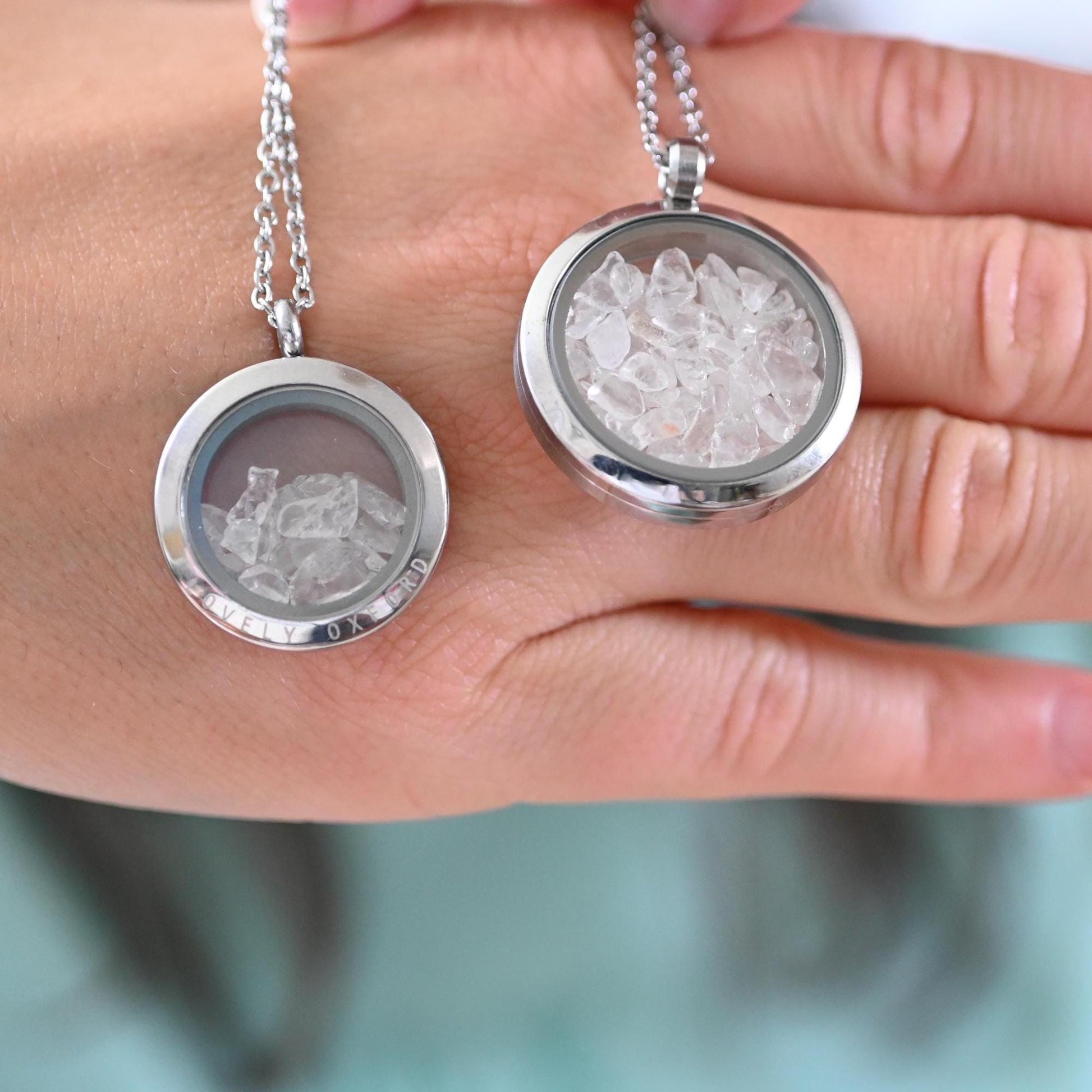Two silver locket necklaces with engraved designs held in a hand against a blurred background.